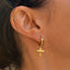 A close-up of a person's ear wearing a delicate gold huggie hoop earring with a dangling textured bar and small charm, alongside a small gold ear cuff.